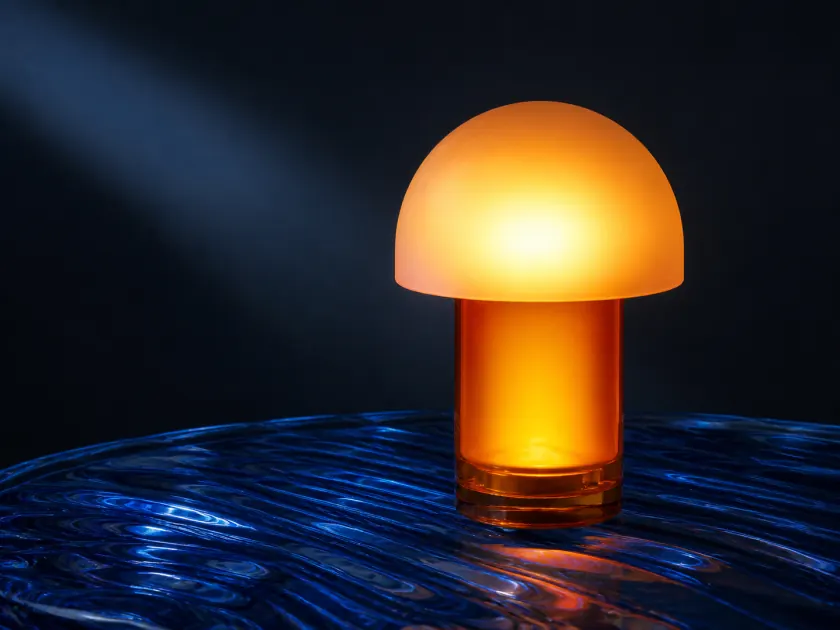 Concept product photograph of a translucent amber table lamp resting on rippled cobalt-blue glass; a warm frosted dome against cool directional light, precise reflections, minimal dark studio, and a premium industrial-design campaign aesthetic.
